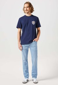 T-shirt in cotone blu navy con maniche corte, con una toppa grafica sul petto sinistro; abbinato a jeans celesti e scarpe grigie.