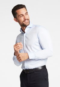 Camicia azzurro chiaro con colletto classico, maniche lunghe e silhouette aderente, indossata con pantaloni scuri e una cintura nera.