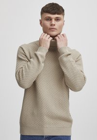 Beige knitted turtleneck sweater with a textured diamond pattern, ribbed cuffs, and hem. Model holds neck with hands.