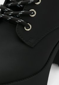 Bota preta de salto alto feita de material matte, com ilhós metálicos redondos e atacadores às riscas pretas e brancas. Design de biqueira arredondada.