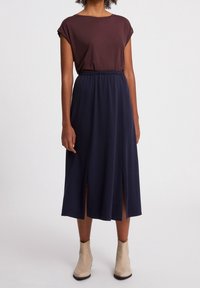 Hnědé přiléhavé tričko s krátkým rukávem v kombinaci s námořnickou midi sukní s dvěma bočními rozparky, doplněné světlými kotníkovými botami.