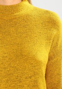 Suéter de punto amarillo con cuello alto, tejido texturizado, patrón acanalado y variaciones sutiles de color.