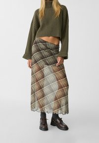 Afgekorte olijfgroene rib gebreide trui gecombineerd met een doorschijnende geruite maxi rok in aardetinten. Model draagt donkere veterschoenen en sokken.