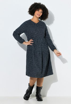 Nainen, jolla on kiharat hiukset, pukeutuneena pitkähihainen polvipituinen tumma kuvioitu mekko ja mustat nilkkurit, seisoo valkoista seinää vasten.