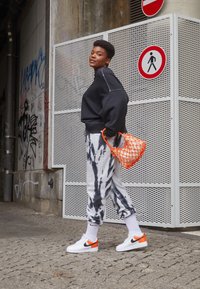 Svart kort sweatshirt, tie-dye mjukisbyxor i grått och svart, vita sneakers med orange detaljer och en orange nätväska. Urban bakgrund.