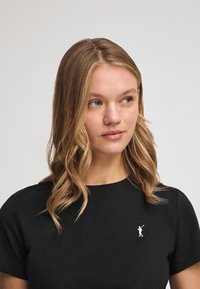 Svart bomull t-shirt med kort ärm, som har en liten vit broderad logotyp på övre vänstra sidan. Slät textur och klassisk passform.