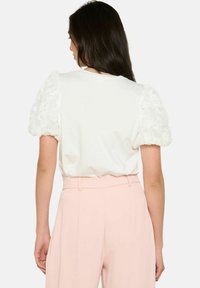 Balta īsā piedkāja tops ar teksturētām, pūkainām mežģīņu piedurknēm. Nēsāts kopā ar augsto jostu gaiši rozā biksēm.