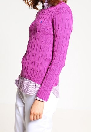 Kvinde iført en lilla kabelstrikket sweater over en lyserød stribet skjorte og hvide bukser, stående sidelæns mod en hvid baggrund.