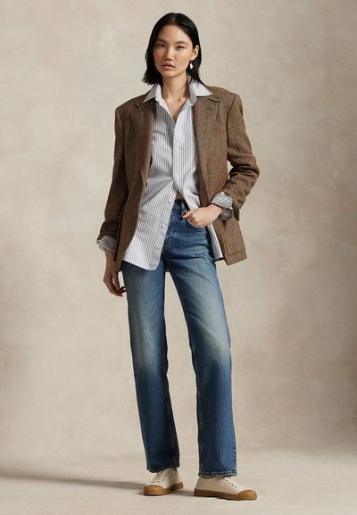 Młoda kobieta w brązowej marynarce, w paskowaną koszulę, niebieskich jeansach i białych trampkach, stoi z ręką w kieszeni na jednolitym tle.