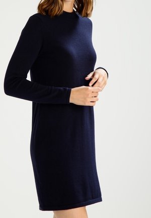 Femme portant une robe ajustée bleu marine à manches longues, arrivant au niveau des genoux, sur un fond clair uni.