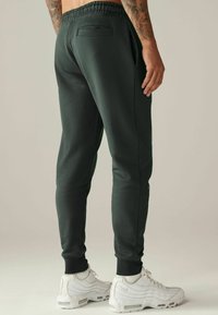 Dunkelgrüne Jogginghosen mit einem elastischen Bund, Bündchen an den Knöcheln und einer Gesäßtasche, kombiniert mit weißen Turnschuhen.