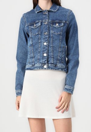 Femme portant une veste en jean bleue avec des boutons argentés et une jupe blanche côtelée, debout devant un fond clair uni.