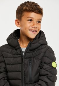 Chaqueta negra acolchada con capucha, bolsillo frontal con cremallera y parche redondo de logo verde en el brazo izquierdo. Tejido suave y brillante con diseño acolchado.