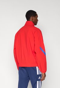 Red windbreaker jacket with blue accents, elastic cuffs, and waistband, featuring a standing collar and a smooth fabric texture.