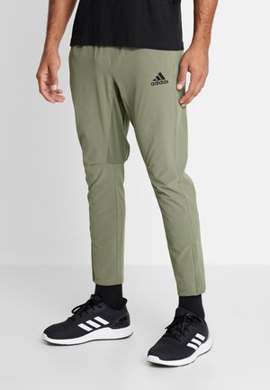 Olivgrüne Sporthosen mit schmaler Passform, die ein schwarzes Adidas-Logo auf dem linken Oberschenkel und texturierte seitliche Akzente aufweisen. Kombiniert mit schwarzen Turnschuhen.