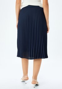 Marineblauwe geplooide rok met een gladde textuur, knielange pasvorm en elastische tailleband, gecombineerd met witte hakken.
