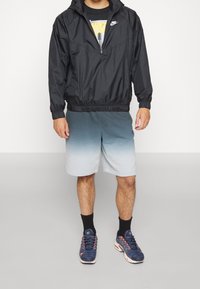 Mustan tuulenpitävän takin, jossa on logo, harmaan sävyttävät shortsit ja siniset lenkkarit. Takissa on säädettävä huppu ja joustava vyötärö.