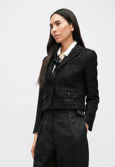 Fekete, texturált anyagból készült, rövidített blézer, két elülső zsebbel, egygombos rögzítéssel és testhezálló szabással, magas derekú fekete nadrág felett.