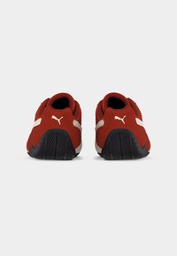 Κόκκινα αθλητικά παπούτσια Puma με λευκές ρίγες και λογότυπο ορατό στο πίσω μέρος, μαύρες υφασμάτινες σόλες, τοποθετημένα σε απλό λευκό φόντο.