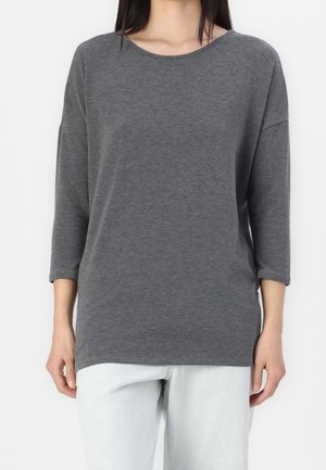 Vrouw draagt een effen donkergrijs shirt met lange mouwen en een ronde hals, gecombineerd met lichtgewassen spijkerbroek, zichtbaar van schouders tot heupen.