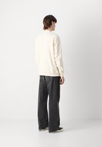 Sudadera crema con puños acanalados, combinada con unos pantalones vaqueros negros de corte suelto, vista desde atrás contra un fondo liso.