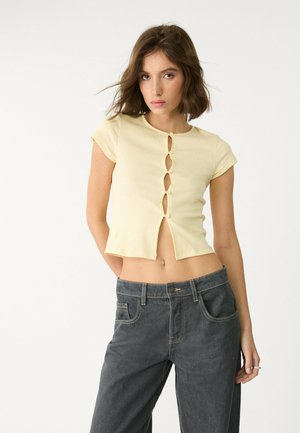 Yellow ribbed crop top with short sleeves and five button detailing down the center. Paired with high-waisted dark gray jeans.