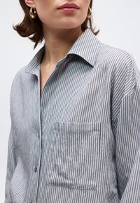 Γυναίκα που φορά γκρι μπλούζα μακρυμάνικη με λεπτές κάθετες μαύρες ρίγες, ορατή τσέπη στο στήθος και χρυσούς κρίκους σκουλαρίκια, μπροστά σε απλό φόντο.