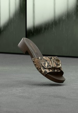 Sandale mule à talon marron avec bride à imprimé serpent, grande boucle dorée et détails cloutés sur fond de sol gris.