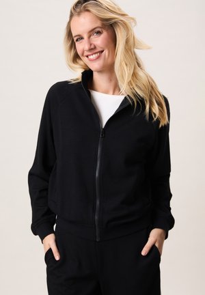 Lachende vrouw met lang blond haar draagt een zwart jack met rits en bijpassende broek, handen casual in de zakken, op een neutrale achtergrond.