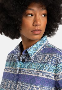 Button-up shirt with a multicolored geometric pattern in blue, green, and purple. Features a collar and textured fabric with button details.