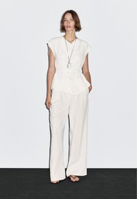 Hvid peplum-top med korte ærmer; parret med flowy, vide bukser. Modellen bærer en pendant halskæde og flade sandaler på et mørkt gulv.