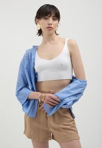 Top corto bianco a coste, camicia azzurro chiaro ricamata, pantaloni corti marroni e bracciali dorati. Presenta una texture morbida e un design a strati rilassato.
