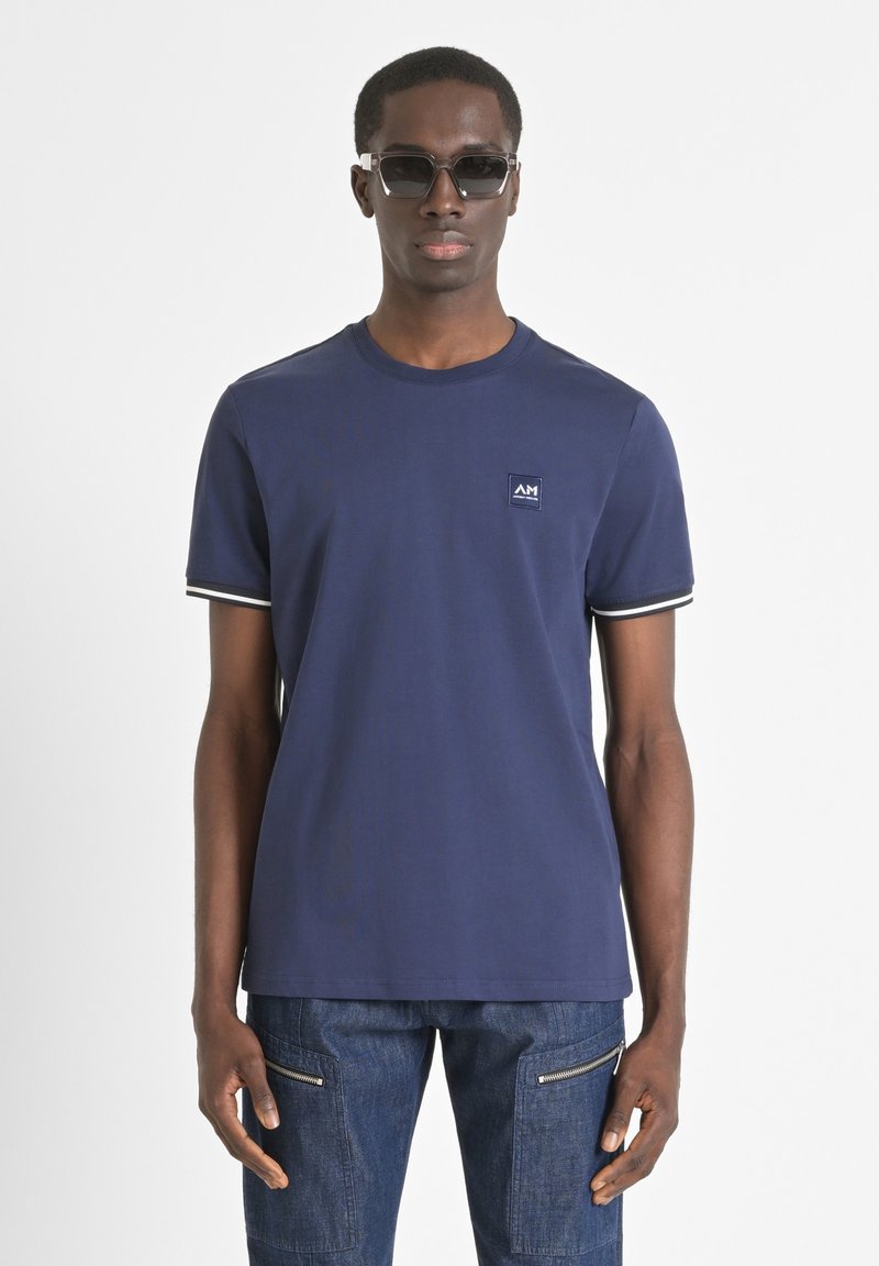 Camiseta azul marino con cuello redondo, de mangas cortas y un diseño ajustado. Cuenta con un pequeño parche de logo y acentos de rayas en las mangas.