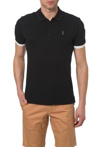 Polo shirt noir à manches courtes, avec un col à trois boutons et des poignets blancs. Porté avec un short beige, détails de logo minimaux visibles.