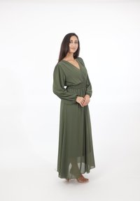 Vrouw staat glimlachend in een olijfgroene maxi-jurk met lange mouwen, een gedefinieerde taille en een transparante zoom, draagt transparante sandalen met hoge hakken.