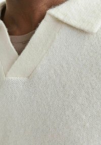 Valge kudumiga pullover V-kaeluse ja kraega. Kangas tundub pehme ja tekstuuriga. Kandmisel helepruuni särgi peal.