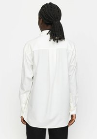 Langärmliges weißes Hemd, durchgehende Knopfleiste hinten, gerader Saum, weicher Stoff, sichtbarer Kragen und Knopfdetails an den Manschetten. Einfaches und klassisches Design.