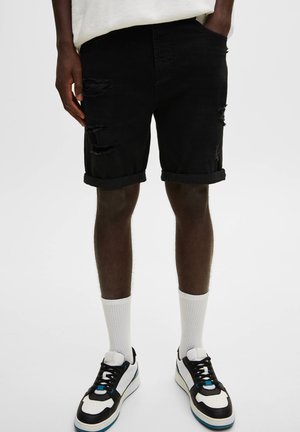 Homme portant un short en jean noir déchiré, des chaussettes blanches mi-mollet et des baskets noir et blanc, debout, les mains partiellement dans les poches.