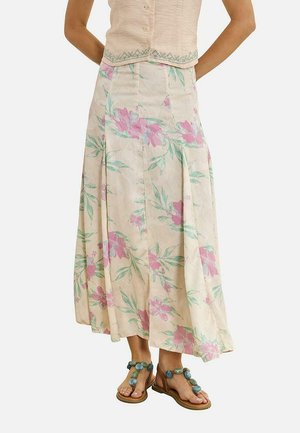 Mujer con falda midi beige de flores rosas, blusa beige bordada y sandalias con grandes adornos de piedra azul.