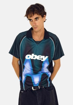 Giovane uomo con polo "Obey" blu e nera con motivo astratto azzurro chiaro, in piedi davanti a uno sfondo chiaro e uniforme.