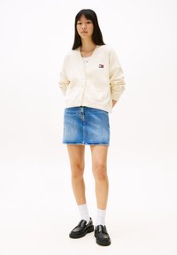 Kremfarget cardigan med knappelukking, V-halsdesign, båret over en lys topp, kombinert med et blått denimskjørt og svarte loafers.