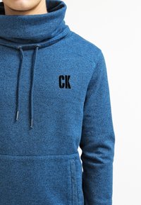 Sudadera azul con un alto cuello, cordón y bolsillo delantero. Presenta el logo bordado en negro "CK". Tejido suave y texturizado con un patrón jaspeado.