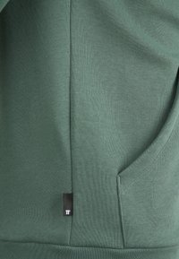 Grüner Hoodie aus weichem, strukturiertem Stoff, mit einer Fronttasche und einem kleinen schwarzen Logetikett. Schlichtes Design mit gemütlichem Aussehen.