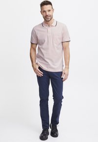 Világos rózsaszín pólóing sötétkék díszítéssel és zsebbel, sötétkék nadrággal és fekete cipővel párosítva, egy sima fehér háttér előtt állva.