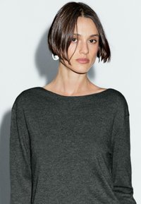 Une femme aux cheveux bruns courts, portant un pull gris foncé, regarde directement l'appareil photo devant un fond gris clair.