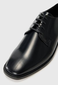 Zapato de vestir de cuero negro con un acabado suave, punta redondeada y cordones estándar. Presenta un sutil pespunte a lo largo del borde de la suela.