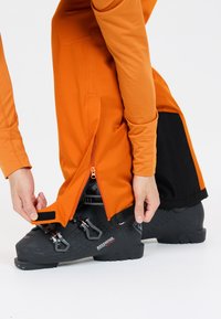 Pantalons de ski orange avec un accent noir, dotés d'une ouverture de jambe zippée et d'une sangle Velcro réglable, positionnés au-dessus de bottes de ski noires.