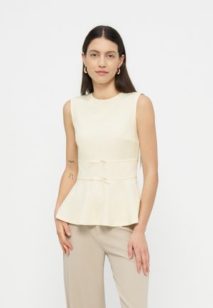 Kvindelig med langt mørkt hår iført guldringøreringe, en ærmeløs cremefarvet peplumtop med bindebånd og beige bukser, stående foran en hvid baggrund.