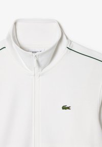 Hvit zip-up jakke med stående krage, grønne akzentstriper og en liten grønn krokodillelogg på brystet. Glatt stofftekstur.
