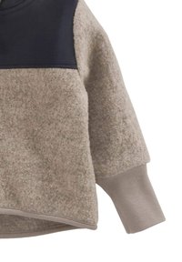 Beige Fleecejacke mit Rippbund am Ärmel, versehen mit einem schwarzen Stoffpanel an der Schulter und einem strukturierten, wollähnlichen Material am Körper.